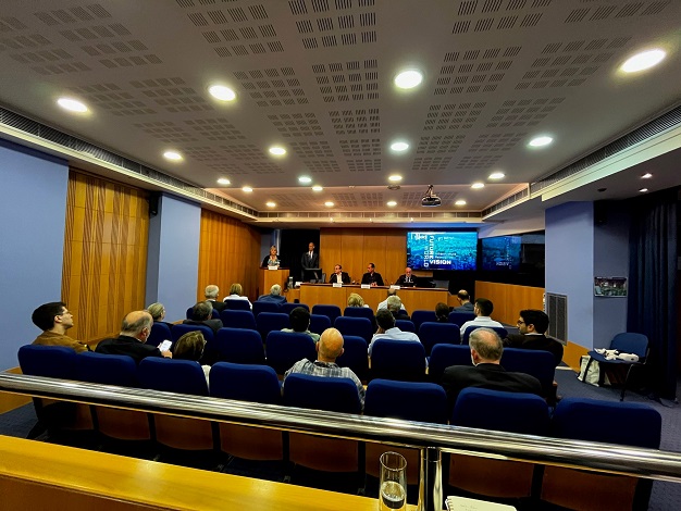 Pictured: Eighteen people sitting in an auditorium watching the presentation given by five presenters in the front of the room next to a screen. 
