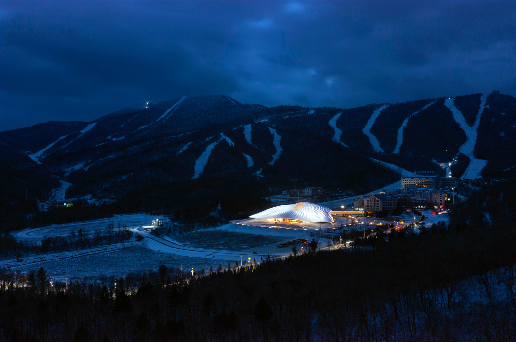 Nighttime image shows an irregular-shaped building lit up at night. The building is surrounded by mountains on one side and forest on the other.