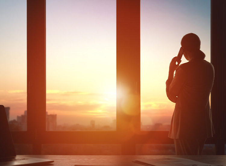 Woman stares out of a window as she talks on the phone. In the background is the outline of a city. 