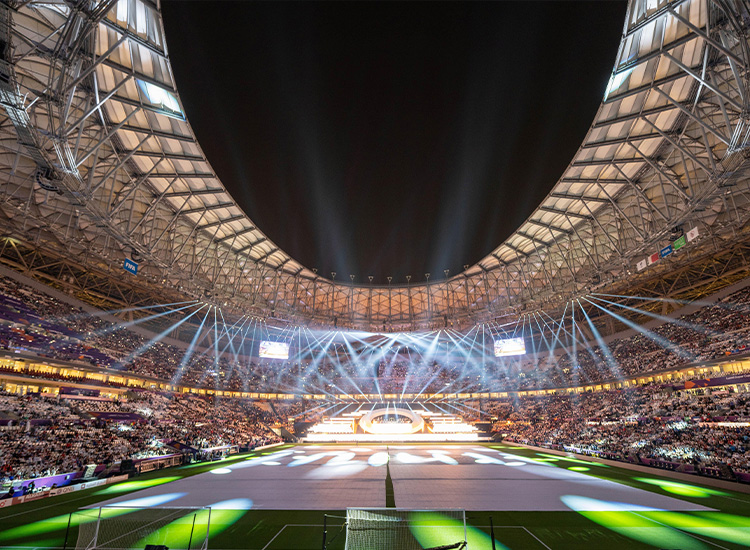 The photograph shows a soccer stadium filled with people and spotlights. 