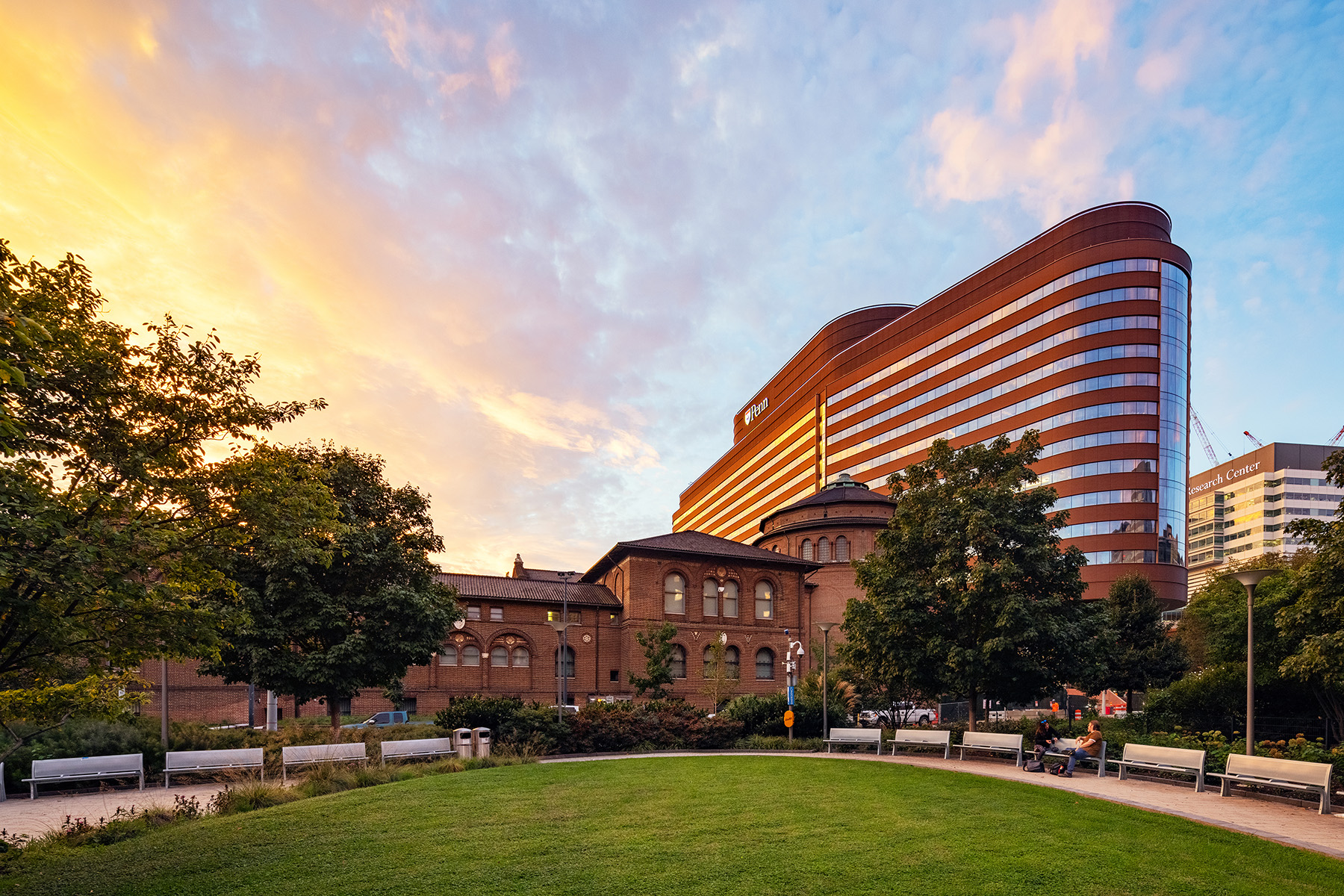Image shows a multistory building with an oblong shape. A greenspace with a path and benches is in the foreground.