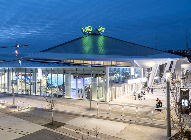 A building with a glass facade has a pointy roof. People walk around outside the building. 