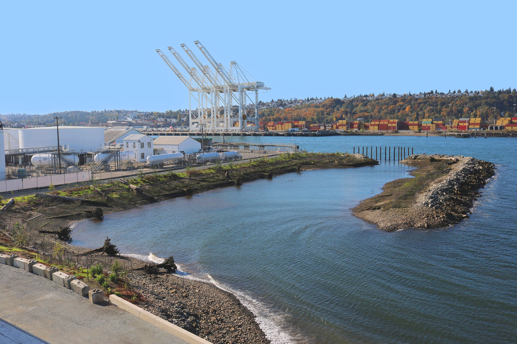 Photograph shows a body of water with industrial buildings on one side and a road on the other. In the water on the right is structure made of earth and rocks with vegetation on it. 