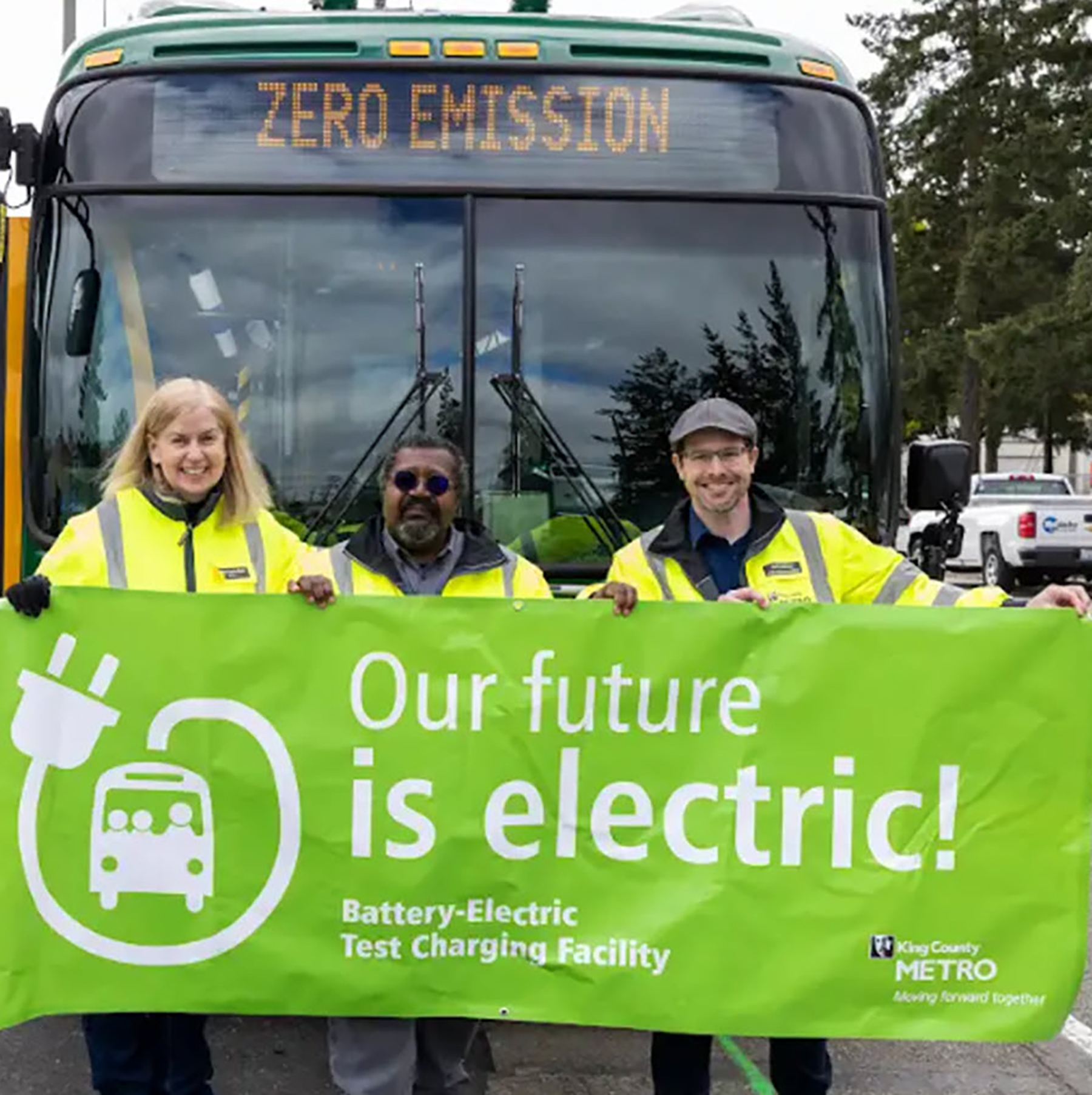 three people smile in front of a bus with a sign promoting electric buses