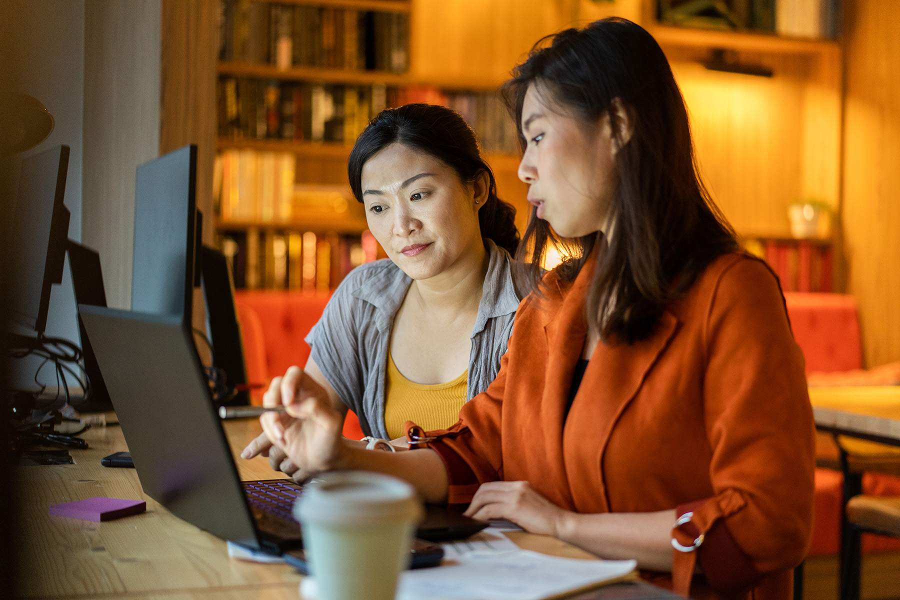 Two Asian women sit at a desk pointing to something on a laptop screen. In the background are blurred bookshelves and in the foreground is a blurry cup of coffee. One woman is wearing a burnt orange shirt and the other a yellow t-shirt with gray sweater. 